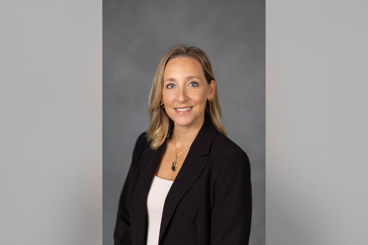 Professional headshot: smiling woman with blonde hair in a black blazer and white top, against a gray backdrop for a bio badge.