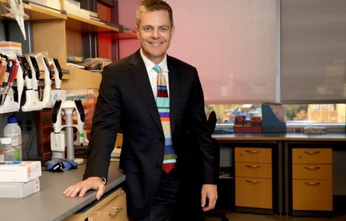 Man in a dark suit standing at a laboratory bench, smiling, with lab equipment and drawers in the background.