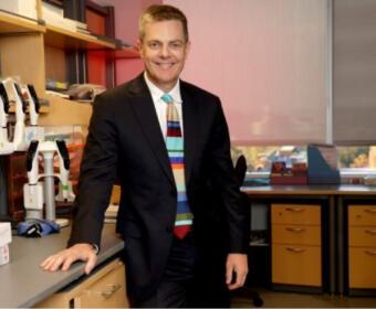 Man in a dark suit standing at a laboratory bench, smiling, with lab equipment and drawers in the background.