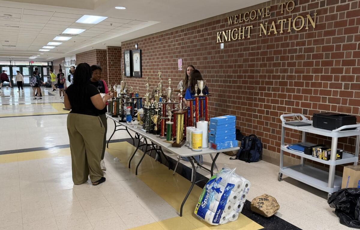 School hallway with a long table full of trophies and supplies under a 'Welcome to Knight Nation' sign on a brick wall