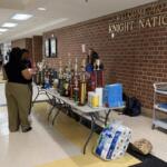 School hallway with a long table full of trophies and supplies under a 'Welcome to Knight Nation' sign on a brick wall