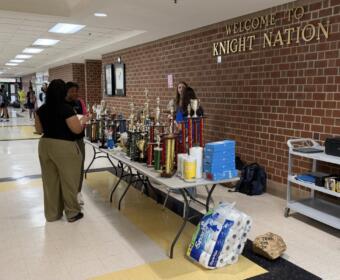 School hallway with a long table full of trophies and supplies under a 'Welcome to Knight Nation' sign on a brick wall