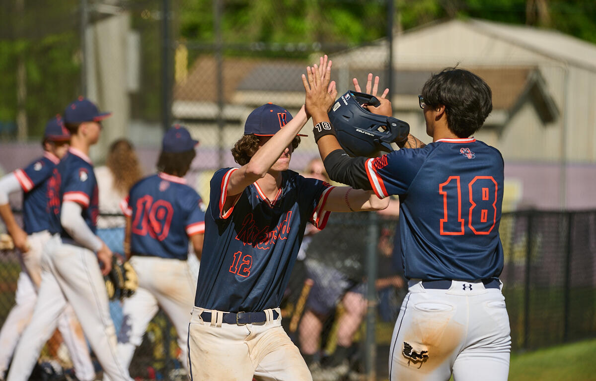 Two baseball players in navy uniforms celebrate with a high-five on the field, near the dugout fence under a sunny sky.