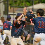 Two baseball players in navy uniforms celebrate with a high-five on the field, near the dugout fence under a sunny sky.