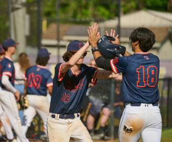Two baseball players in navy uniforms celebrate with a high-five on the field, near the dugout fence under a sunny sky.