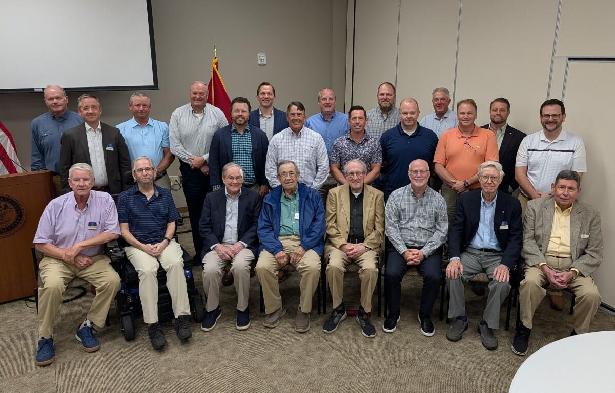 Group of men posing for a group photo in a conference room, with a US flag and podium visible to the left and two-row arrangement of attendees.