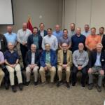Group of men posing for a group photo in a conference room, with a US flag and podium visible to the left and two-row arrangement of attendees.
