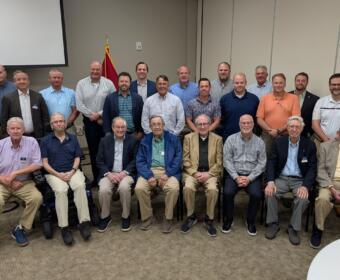 Group of men posing for a group photo in a conference room, with a US flag and podium visible to the left and two-row arrangement of attendees.