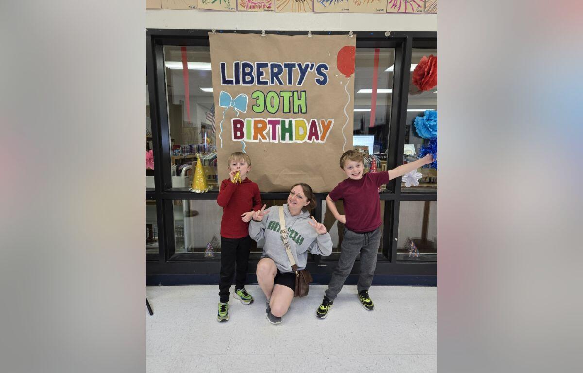 Three people pose in front of a large brown banner reading "LIBERTY'S 30TH BIRTHDAY" with balloons and party decorations in a classroom or hallway setting.