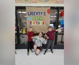 Three people pose in front of a large brown banner reading "LIBERTY'S 30TH BIRTHDAY" with balloons and party decorations in a classroom or hallway setting.