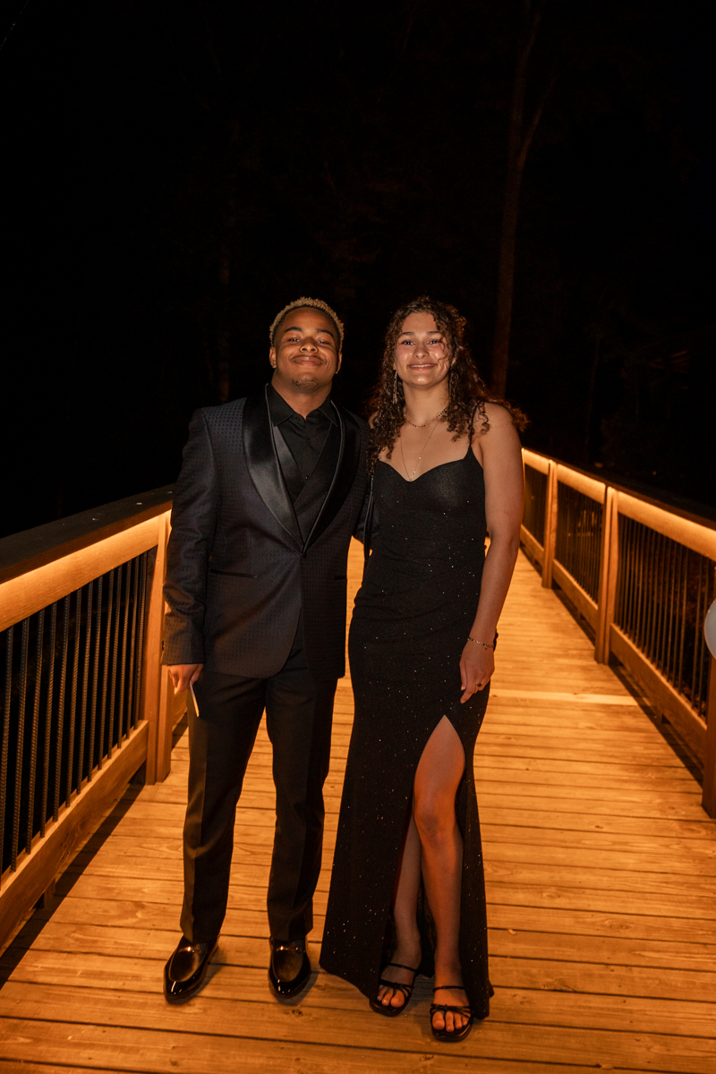 A young man in a black tuxedo and a young woman in a black evening gown pose together on a warmly lit wooden bridge at night .