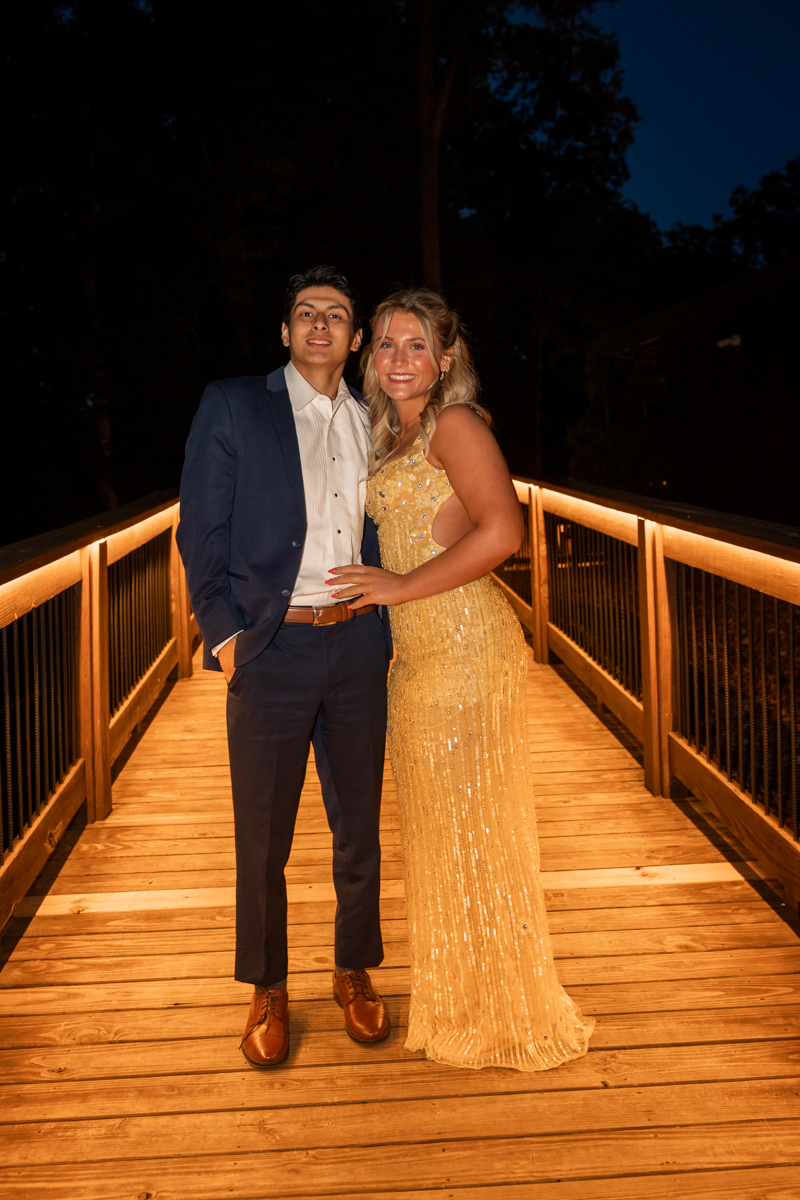 Couple posing on a warmly lit wooden bridge at night; man in a navy suit and brown shoes, woman in a gold sequined gown with blonde hair.