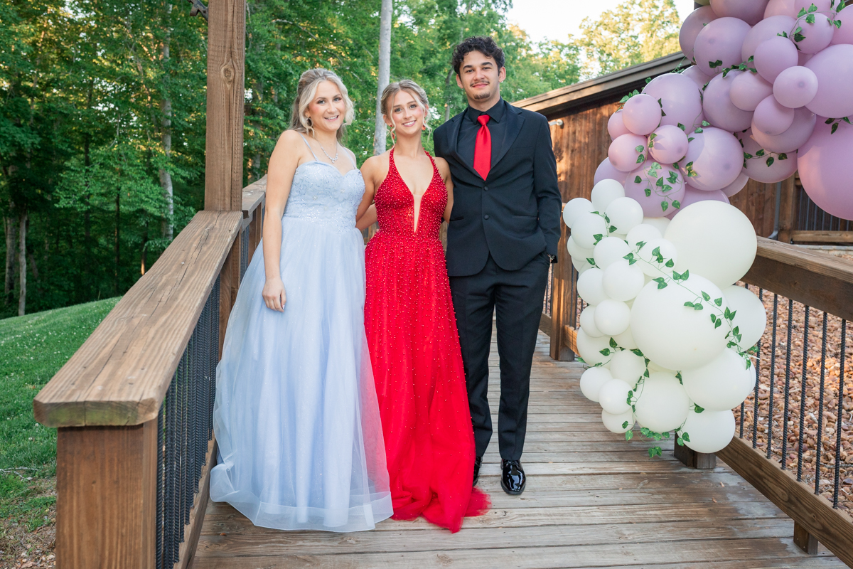 Three young people in formal wear pose on a wooden deck with a pink/white balloon arch to the right, greenery in the background.