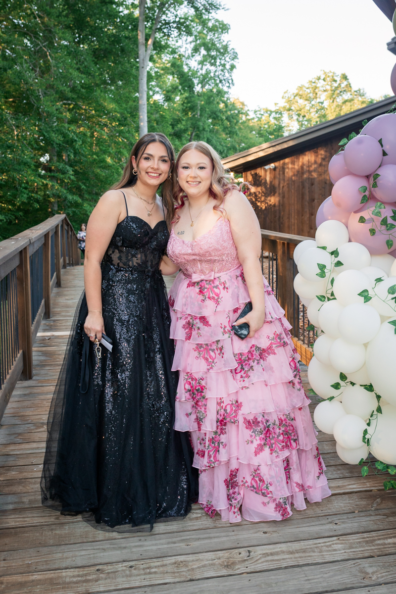 Two women in formal gowns posing on a wooden deck beside a pink-and-white balloon arch; one in black sequin dress, the other in pink floral dress.