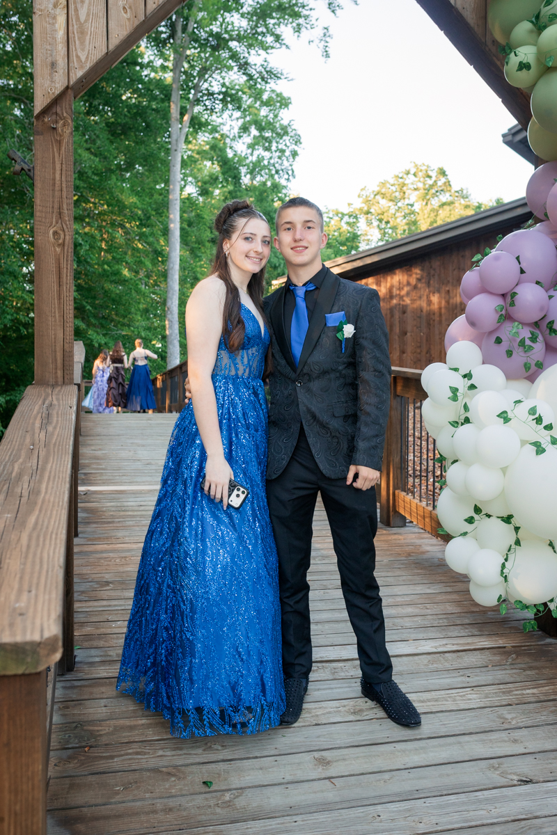 Pair of teens in formal attire posing on a wooden deck, woman in a blue gown holding a phone and man in a dark suit with blue tie, smiling at camera near a pastel balloon arch.