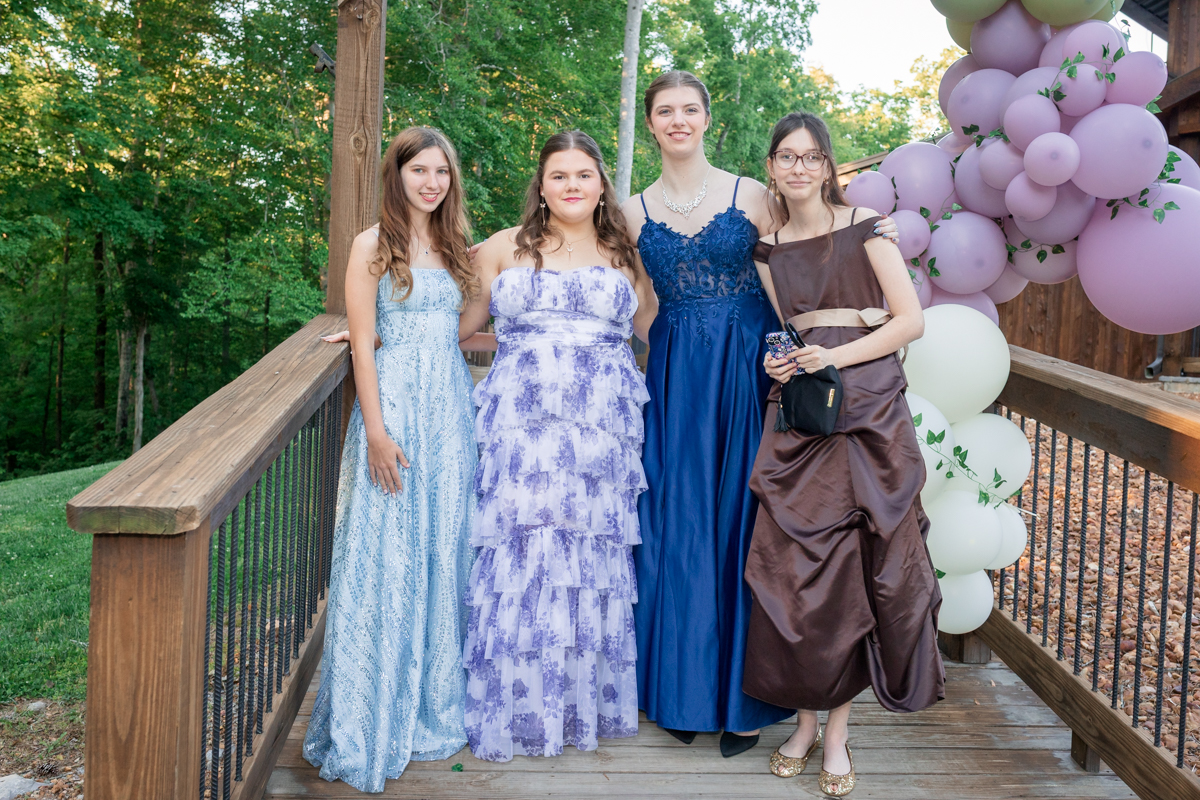 Four young women in formal gowns posing on a wooden deck with purple and white balloons in the background, smiling at the camera.