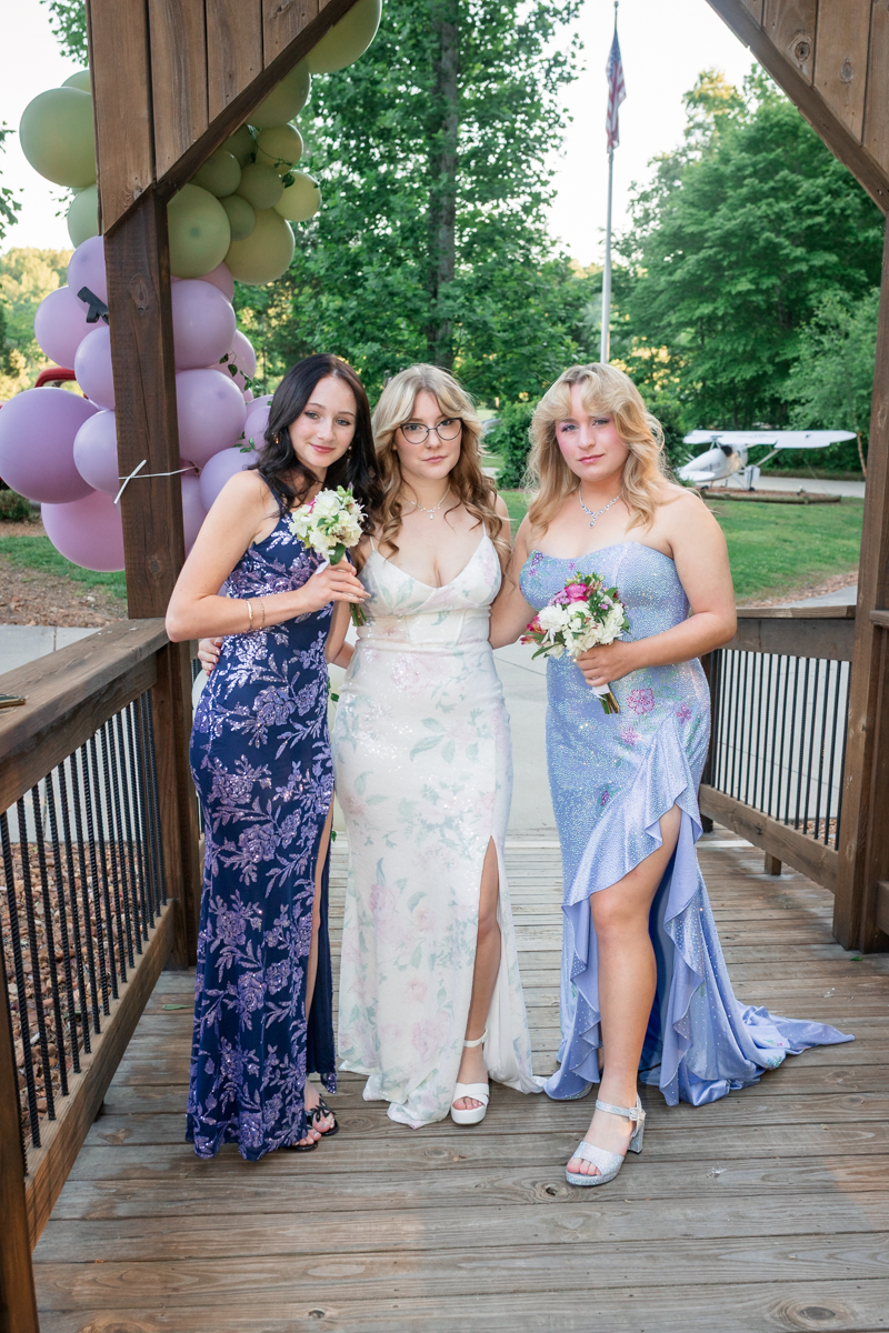 Three women in formal gowns pose together on a wooden bridge with pastel balloons in the background, each holding a small bouquet.