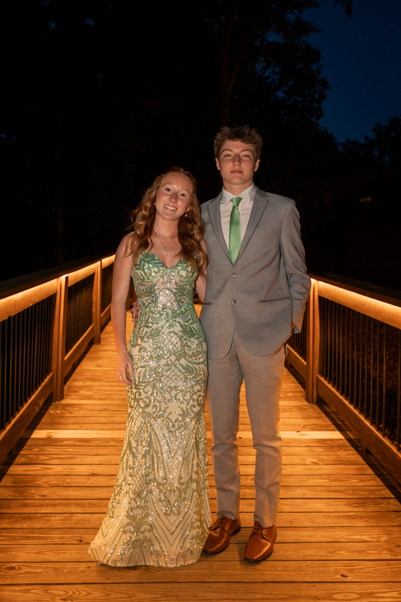 Couple posing on a lit wooden bridge at night; woman in a sequined green gown, man in a gray suit with a green tie.