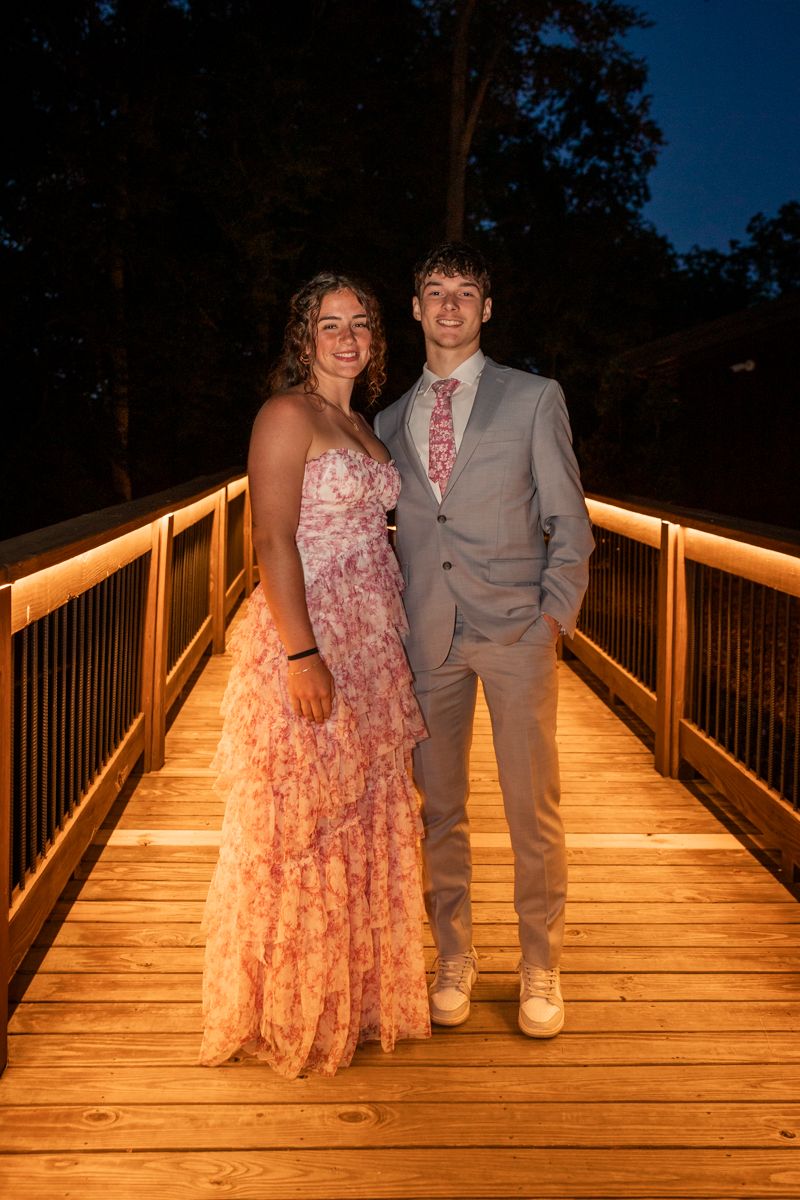 Couple posing on a warmly lit wooden bridge at night; girl in a pink floral dress, boy in a light gray suit and pink tie, both smiling.