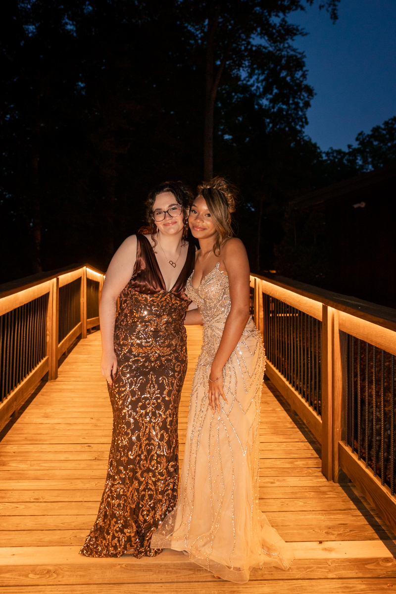 Two women in formal gowns pose together on a warmly lit wooden bridge at night.
