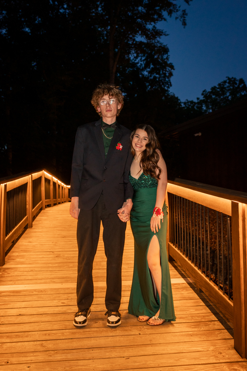 Young couple in formal attire standing on a warmly lit wooden bridge at night, holding hands; man in dark suit, woman in emerald green gown with a slit, smiling at the camera.