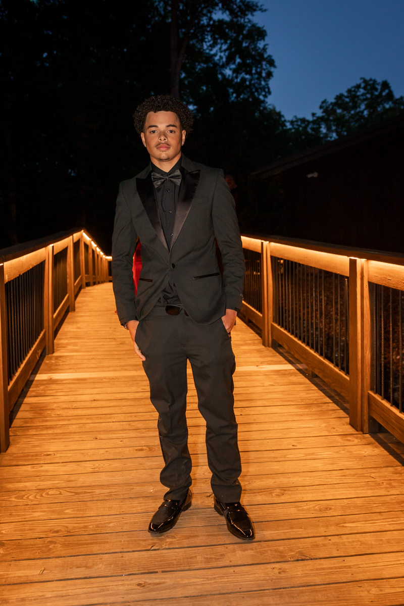 Young man in a black tuxedo with a bow tie stands on a warmly lit wooden bridge at night, hands in pockets.