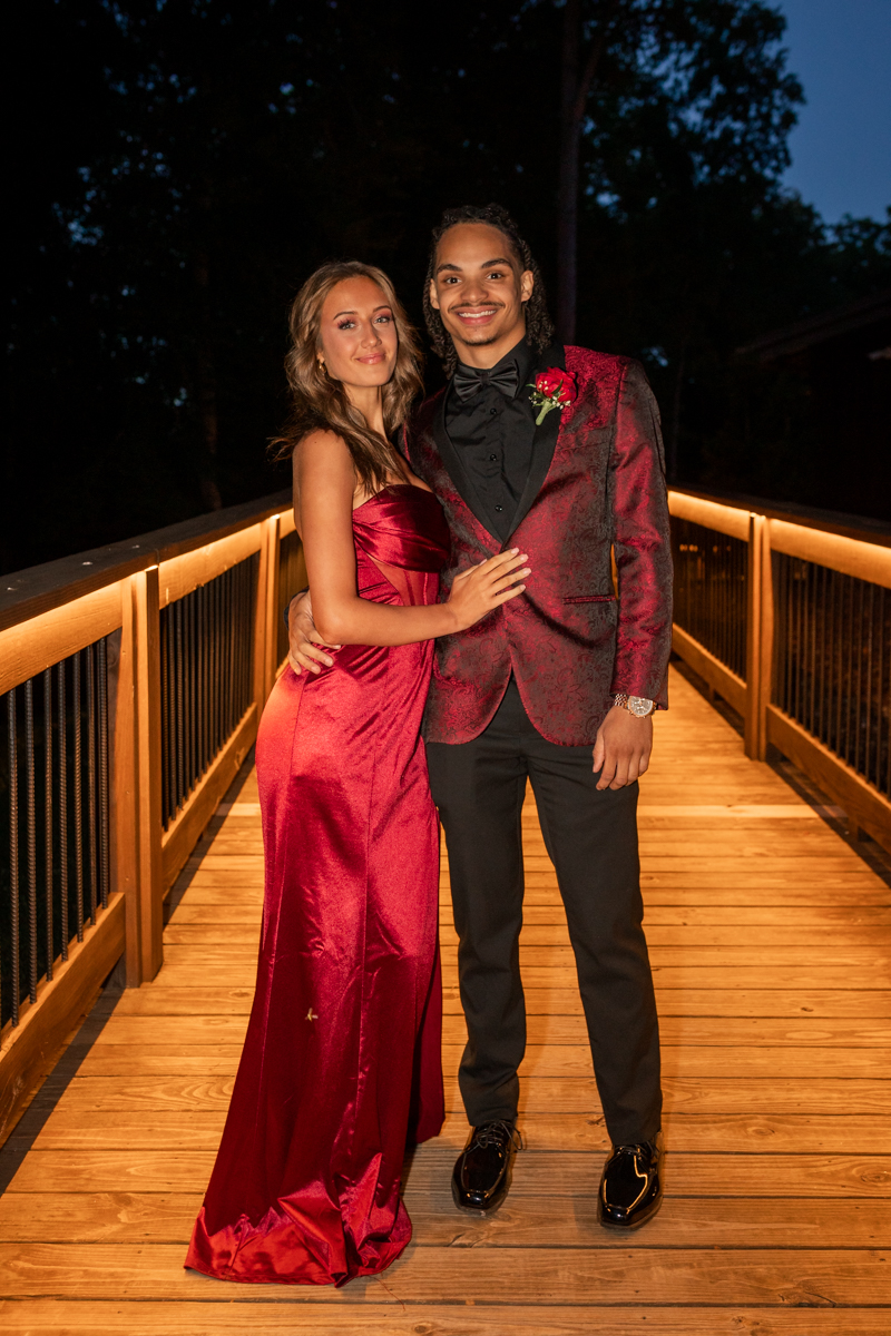 Couple posing on a wooden bridge at night; woman in a red gown and man in a red-tailored suit with a boutonniere smiles at the camera.