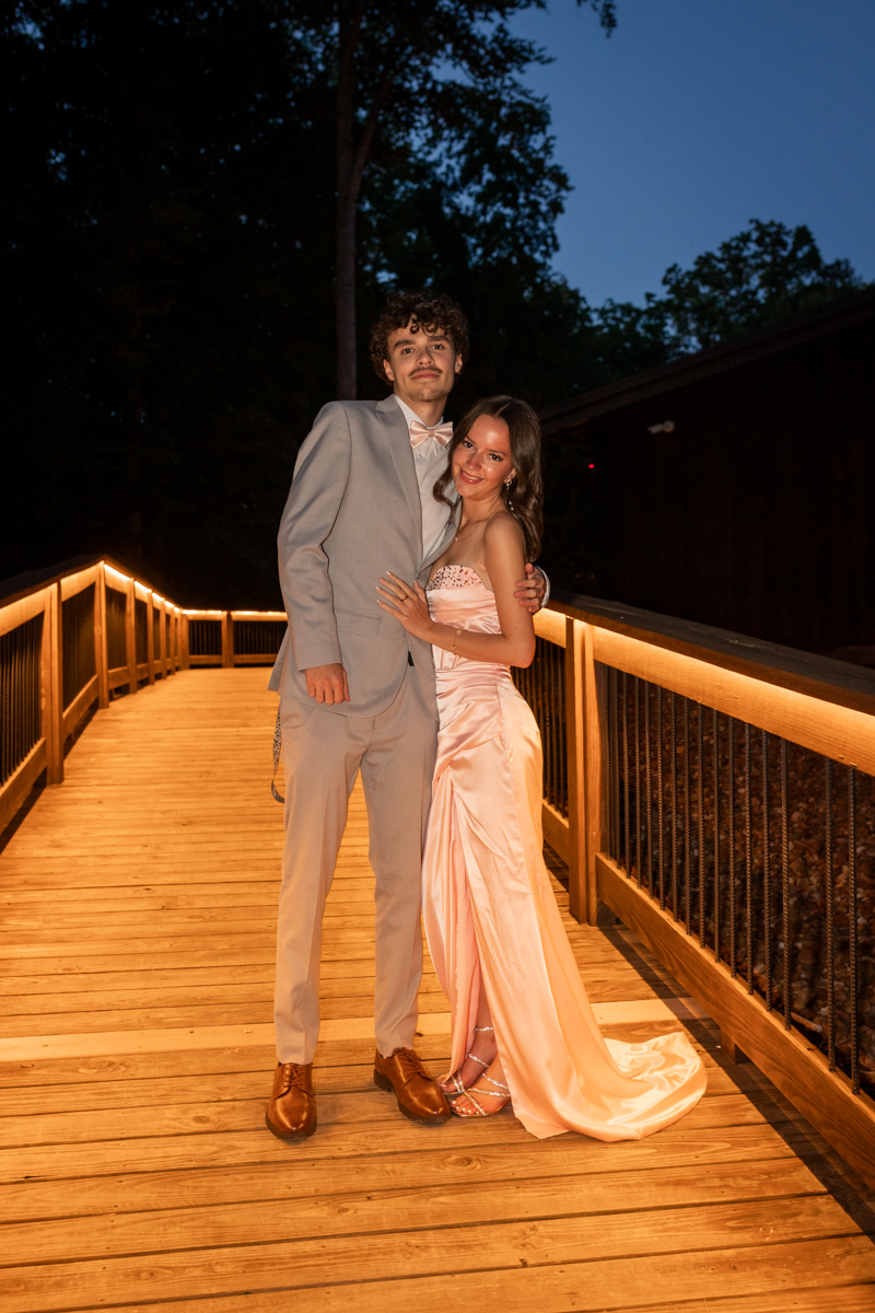 Couple posing on a lit wooden bridge at night; man in a gray suit, woman in a pink gown embrace and smile at the camera.