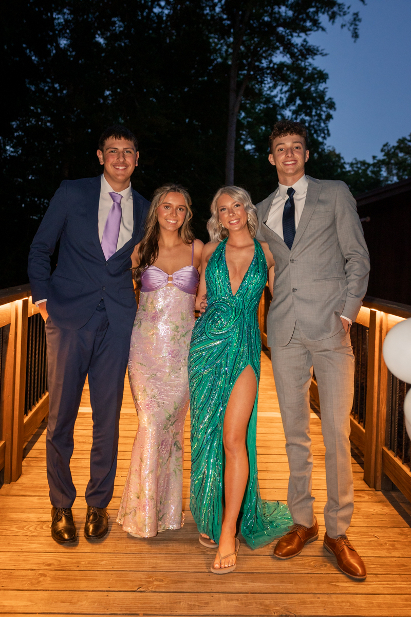 Four teens in formal attire pose together on a lit wooden bridge at night, smiling at the camera on a social event.
