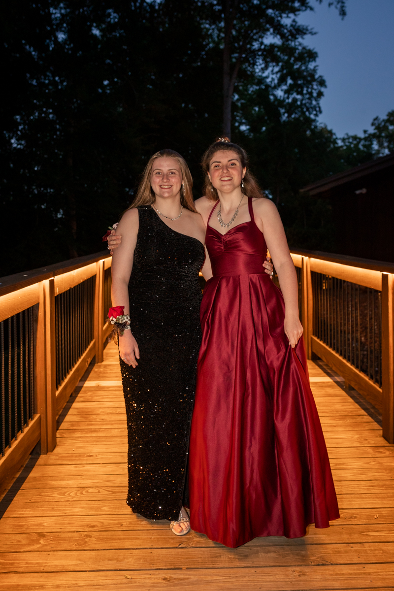 Two young women in formal gowns pose together on a lit wooden bridge at night with trees in the background.