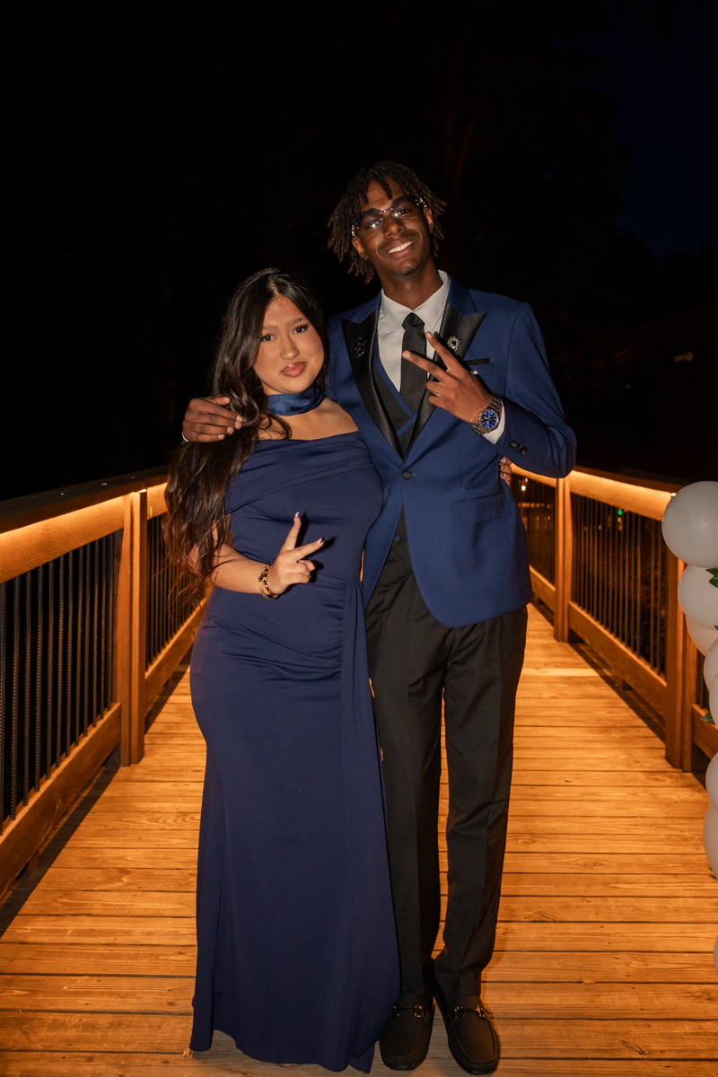 Two young adults in blue formal wear posing together on a warmly lit wooden bridge at night, smiling at the camera.