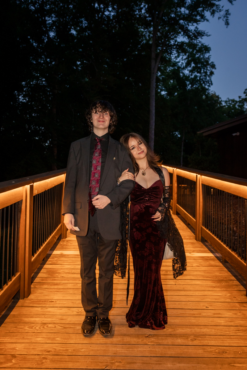 Couple in formal attire walking arm in arm on a lit wooden bridge at dusk or night.