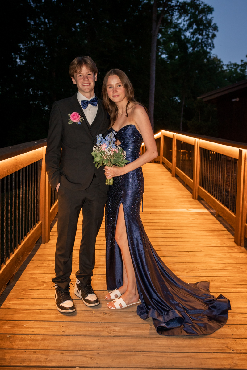 Prom couple posing on a warmly lit wooden bridge; she in a navy sequined gown with a high slit and bouquet, he in a black suit with a blue bow tie and pink boutonniere.