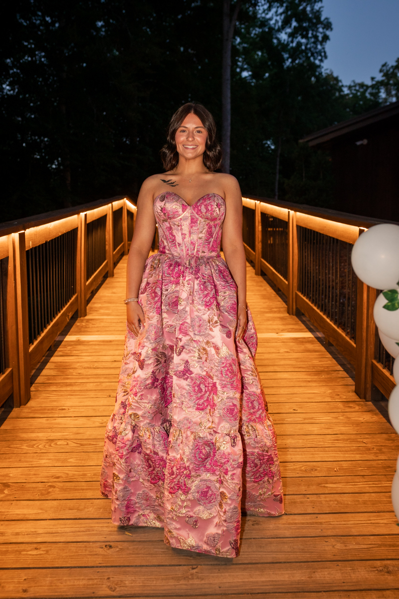 Woman in a pink floral ball gown standing on a lit wooden bridge at dusk, balloons visible on the right.