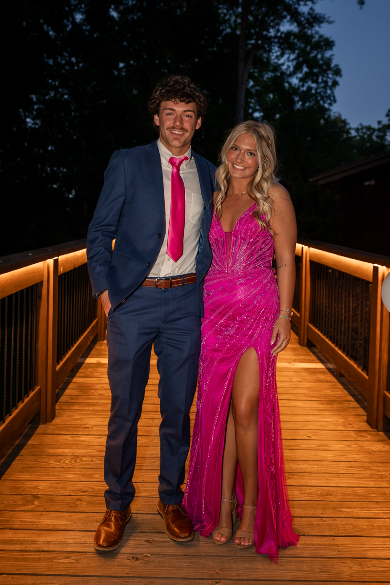 Young couple in formal wear posing on a lit wooden deck at dusk; man in a blue suit with pink tie, woman in a pink sequined gown with slit.