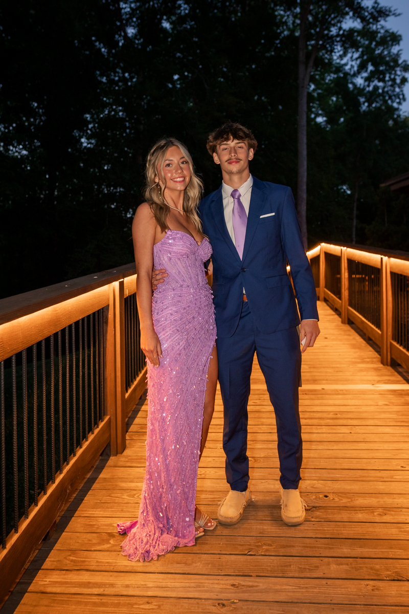 A couple posing on a warmly lit wooden bridge at night, woman in a lavender sequin gown and the man in a blue suit with a lavender tie.