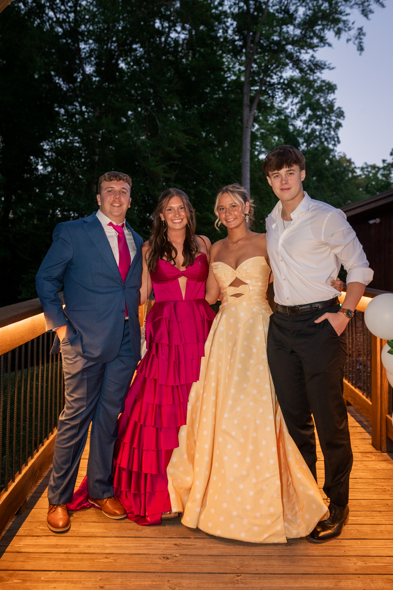 Four friends in formal wear posing on a wooden deck at dusk, with trees in the background.