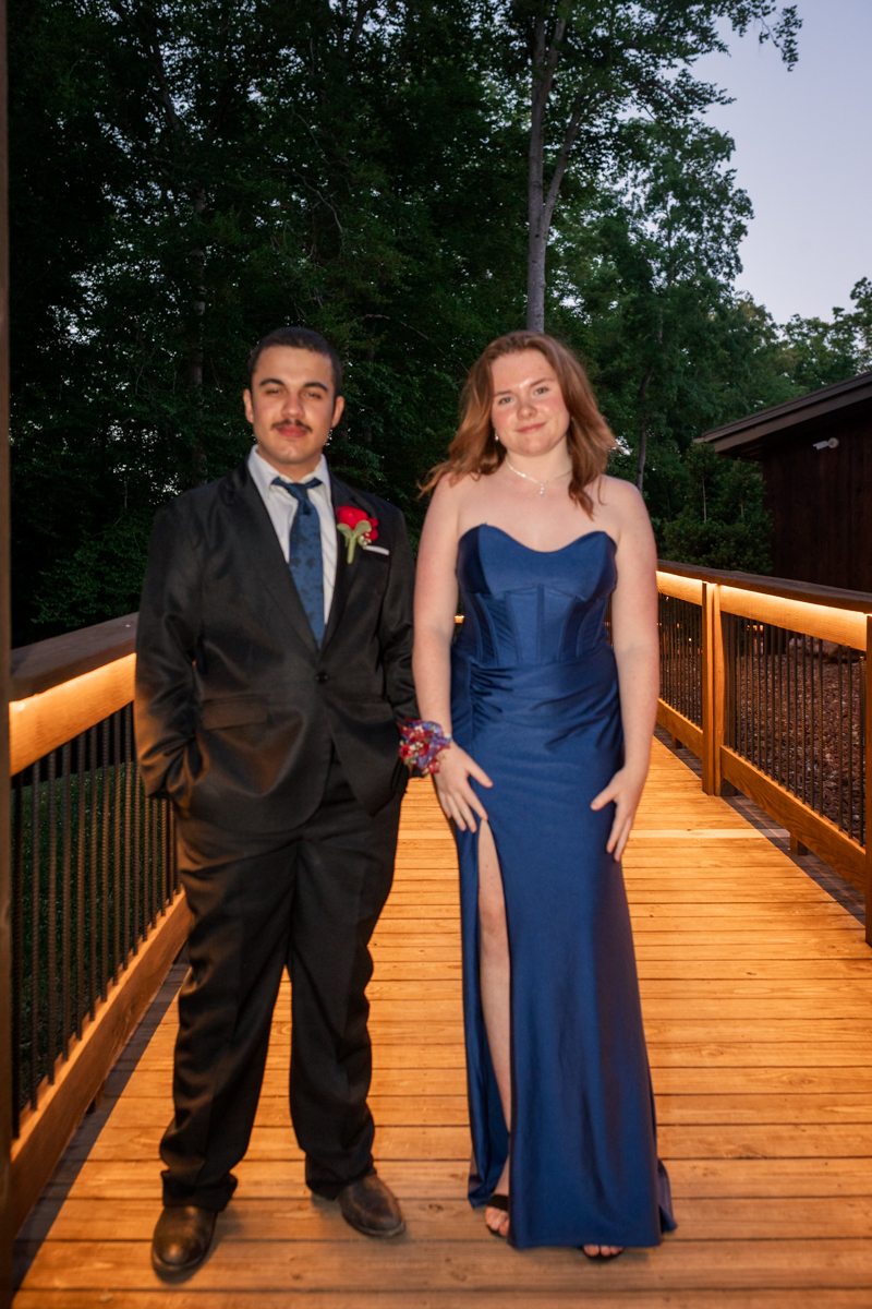 Two young adults pose on a lit wooden bridge outdoors at dusk; man in a black suit with blue tie, woman in a blue strapless gown with a high slit.