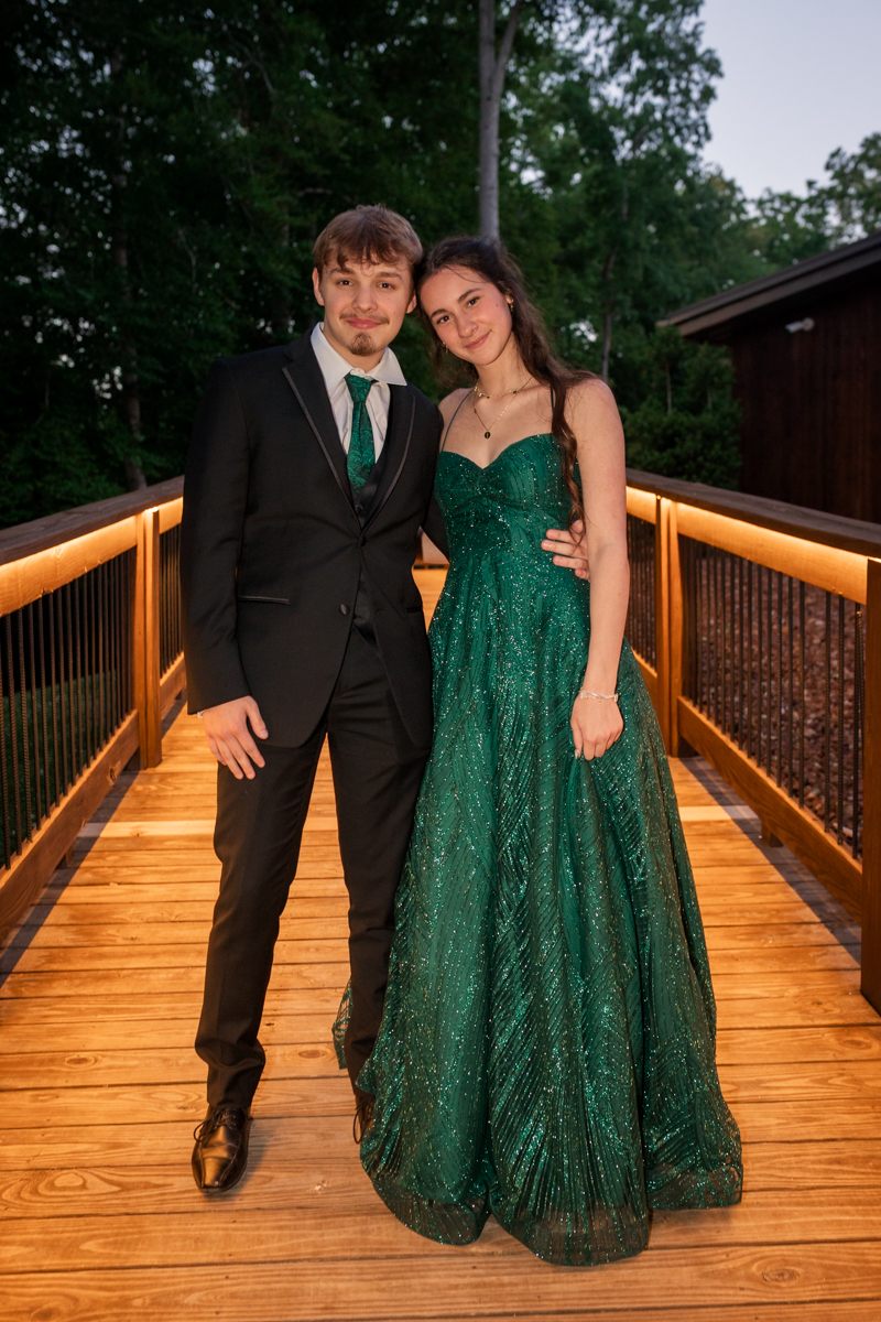 Couple in formal wear posing on a lit wooden bridge: man in a black suit with green tie, woman in a sparkling emerald gown.