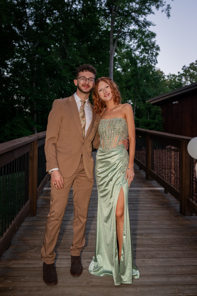 Couple dressed formally posing on a wooden footbridge, man in a tan suit and patterned tie, woman in a light green mermaid gown with a thigh-high slit.