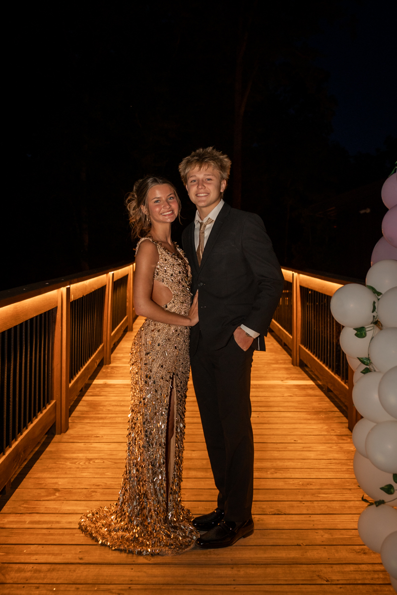 Two teens in formalwear posing arm-in-arm on a warmly lit wooden bridge at night, with white balloons nearby.