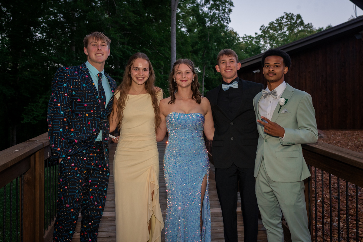Five young people in formal attire posing on a wooden deck outdoors, with trees in the background at dusk.