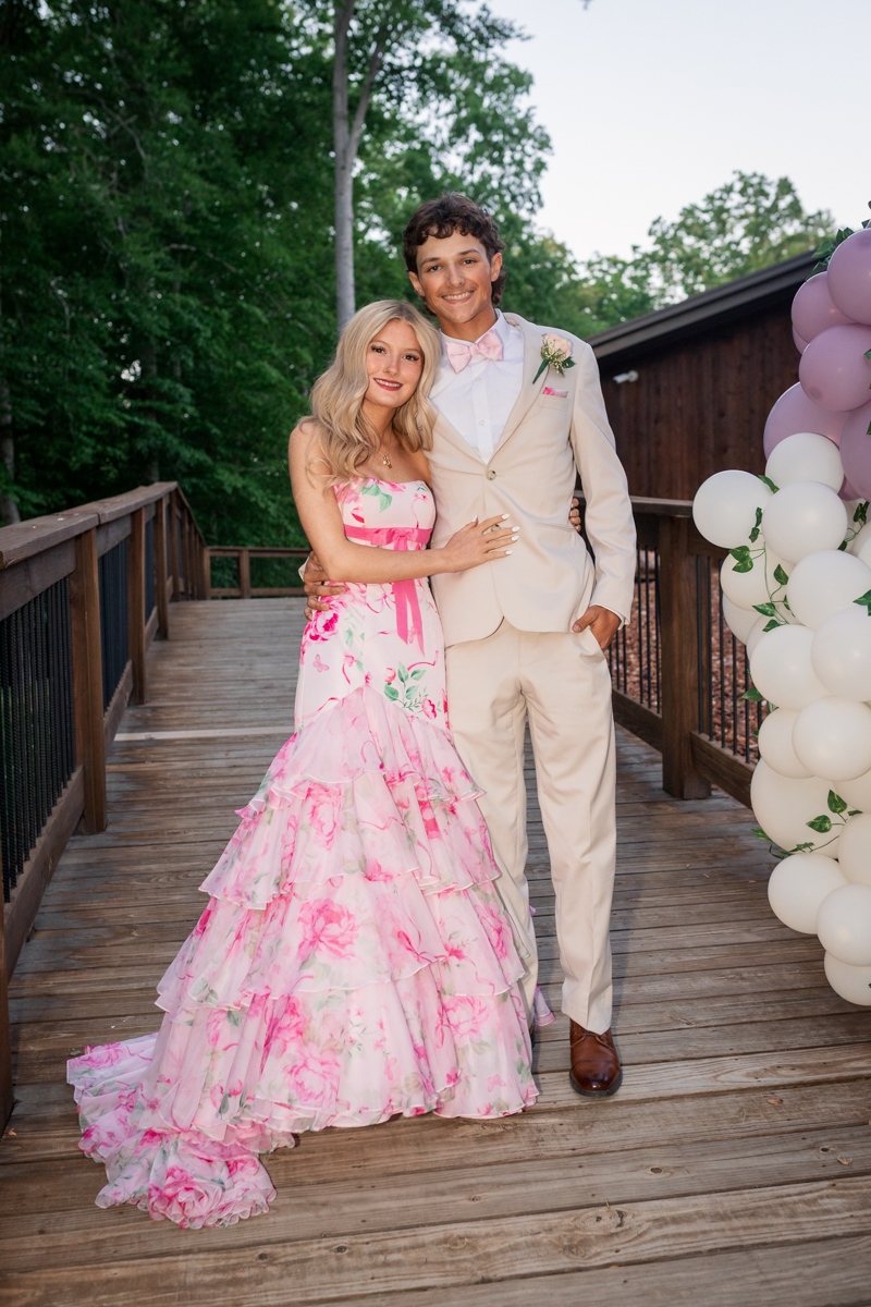 Young couple posing together at an outdoor formal event; woman in a pink floral gown and man in a beige suit with a pink bow tie, embracing on a wooden deck with balloons nearby.