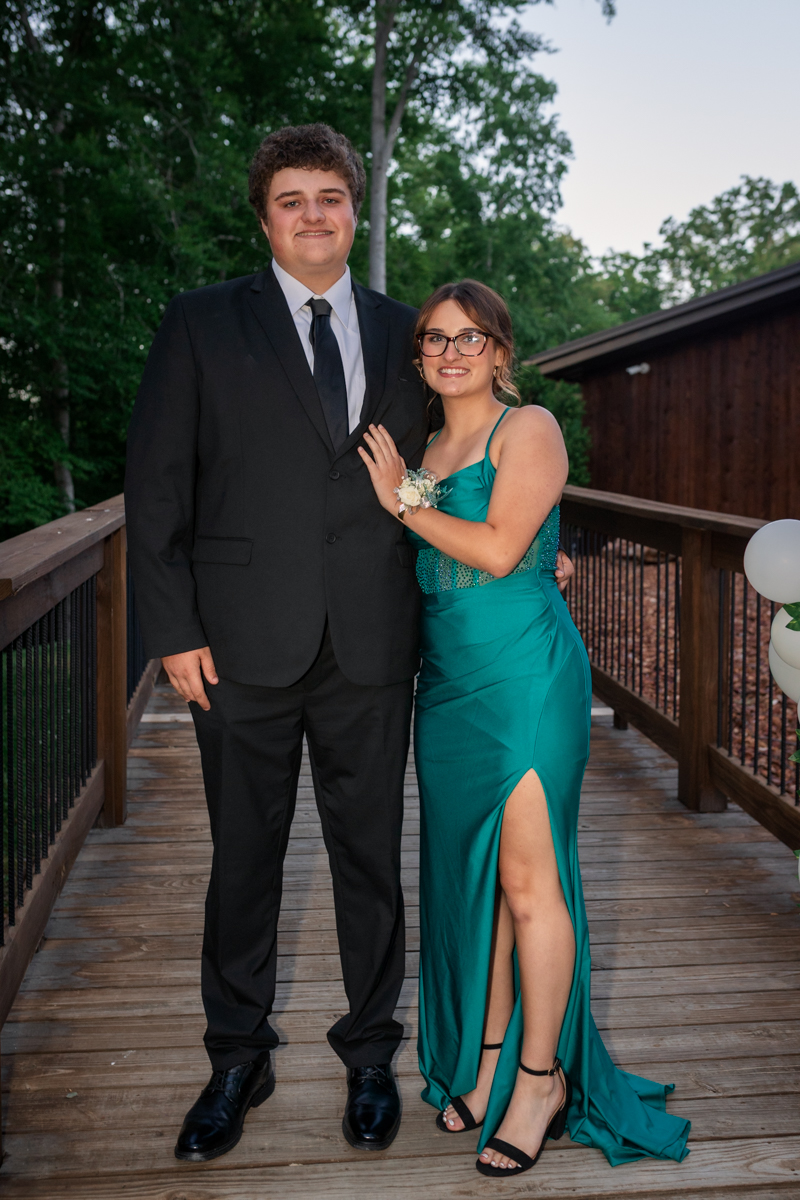 Couple in formal attire posing on a wooden deck outdoors; man in a black suit and tie, woman in a teal gown with a thigh-high slit and a corsage on her wrist.