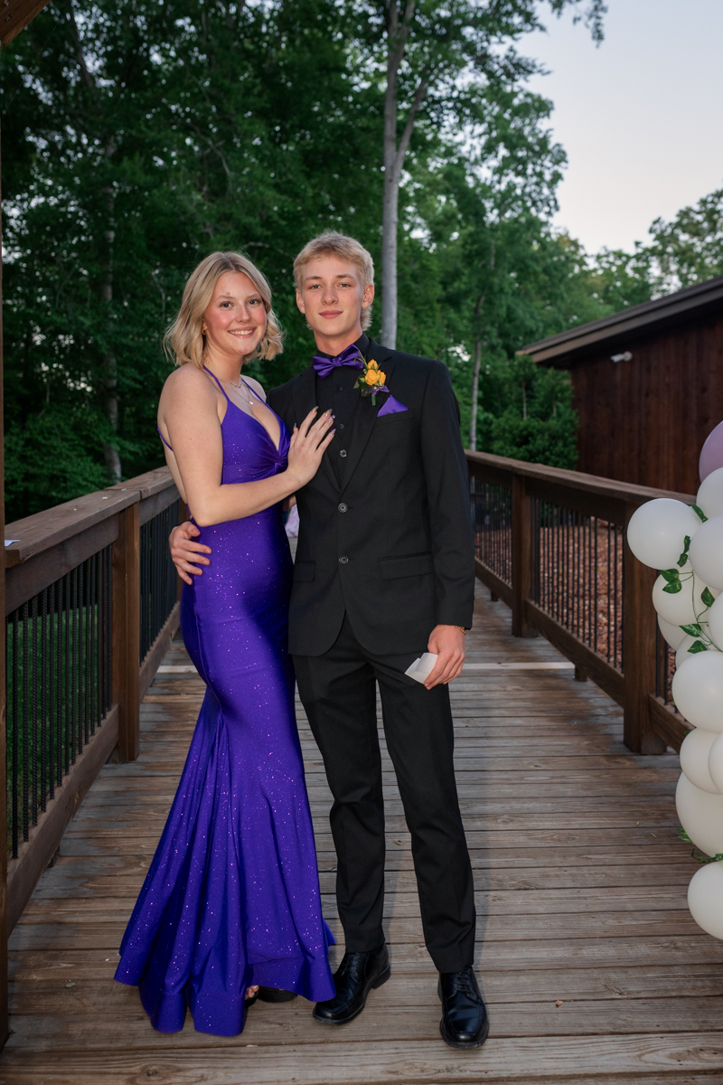 Two teens pose on a wooden bridge; girl in a purple sparkly gown and boy in a black suit with purple bow tie and boutonniere.