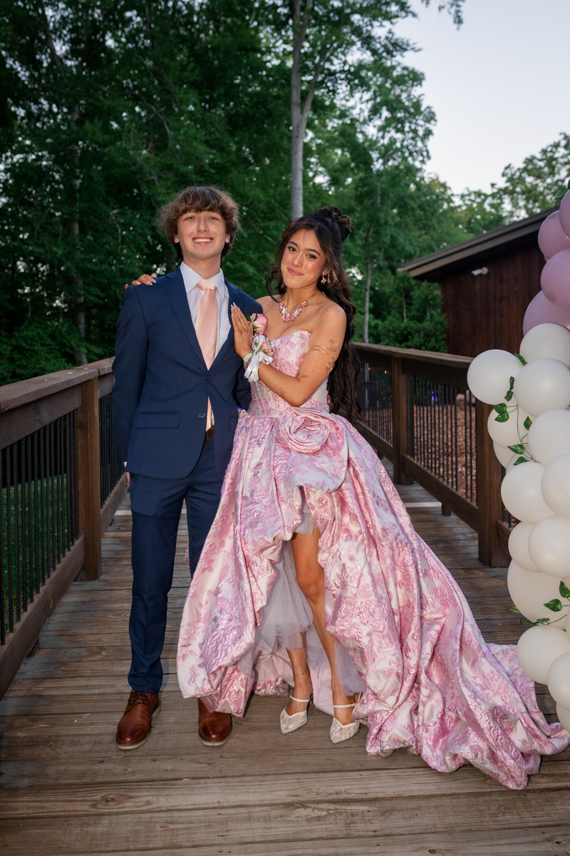 Young couple in formal wear posing on a wooden deck with white and pink balloons; girl in a pink strapless ball gown and the boy in a navy suit.