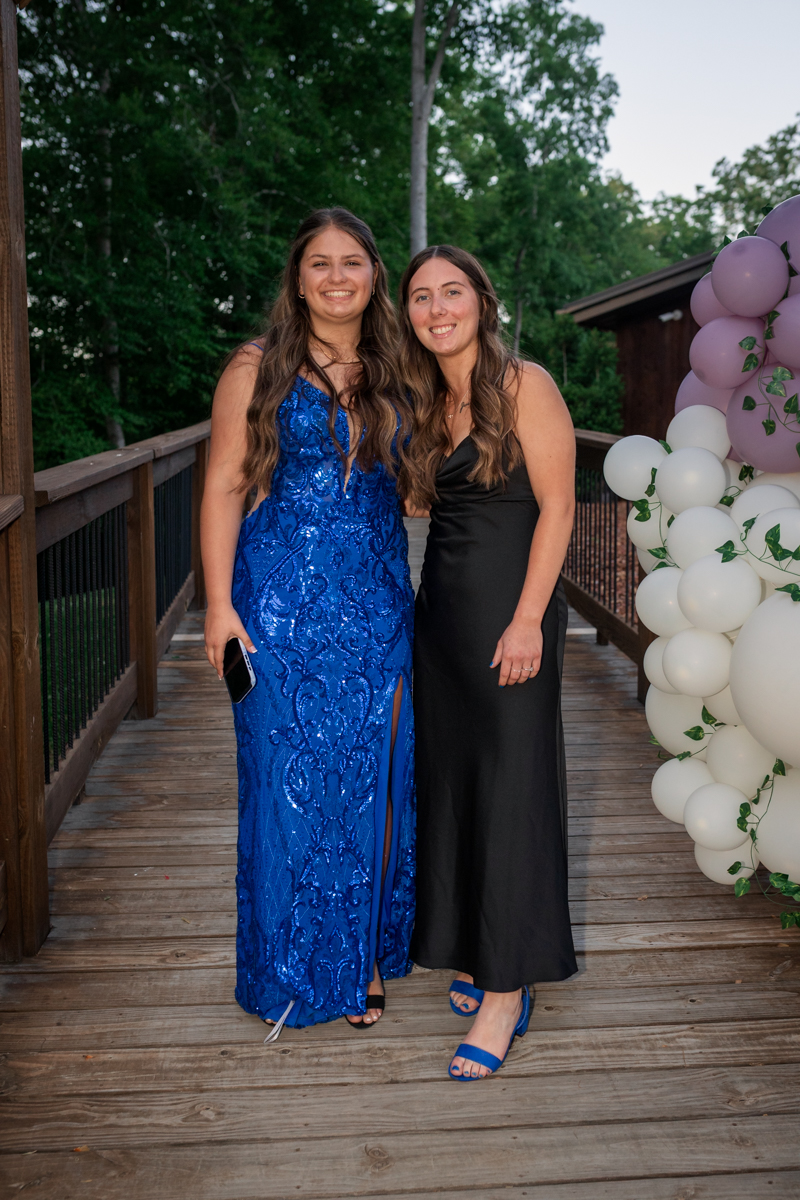 Two young women in formal gowns pose together on a wooden deck, smiling, with a purple–white balloon arch nearby. One holds a phone in her hand.