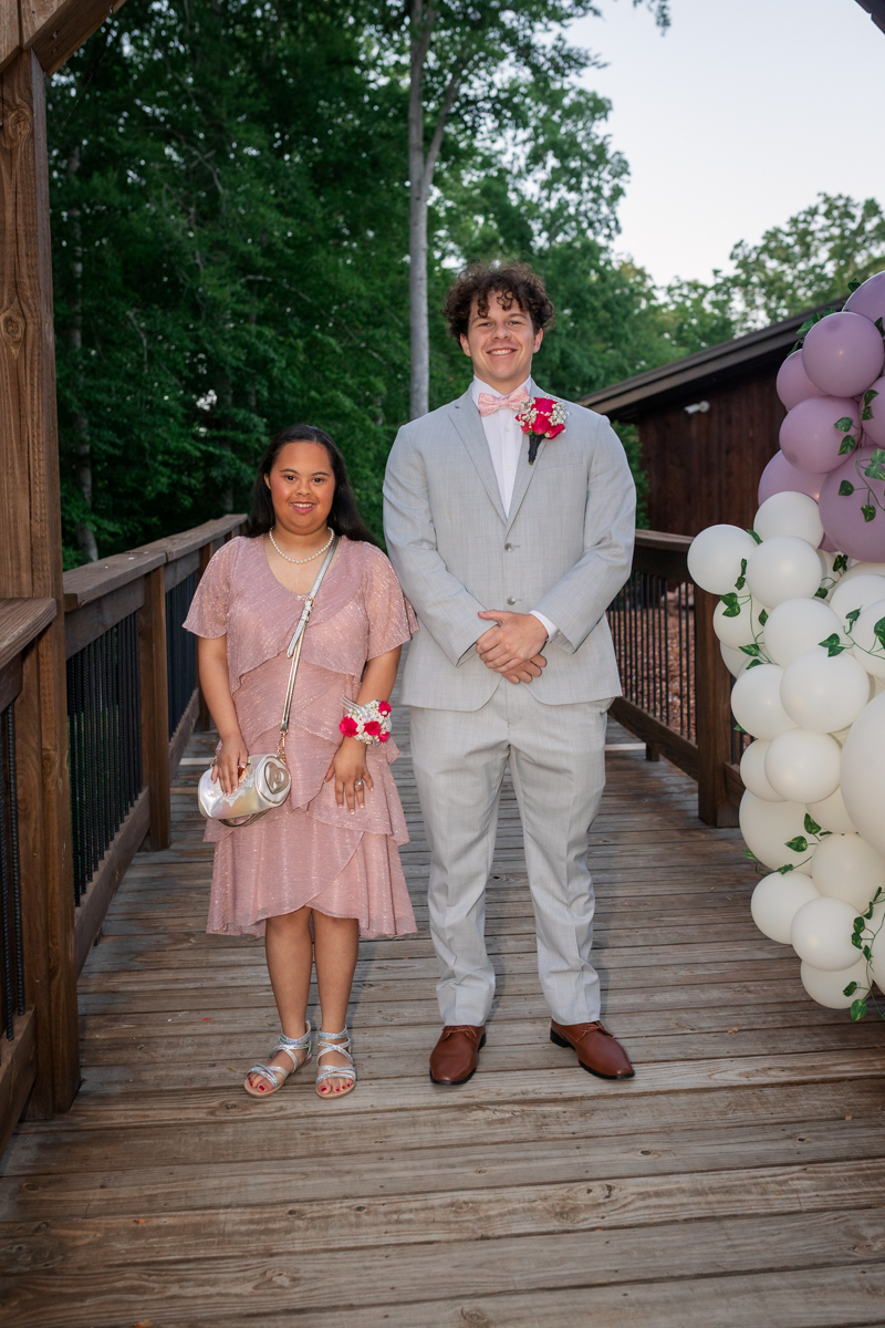 Two teens pose for a photo on a wooden walkway; the girl wears a pink dress with a corsage and the boy wears a light gray suit with a boutonniere, next to a balloon arch outdoors.