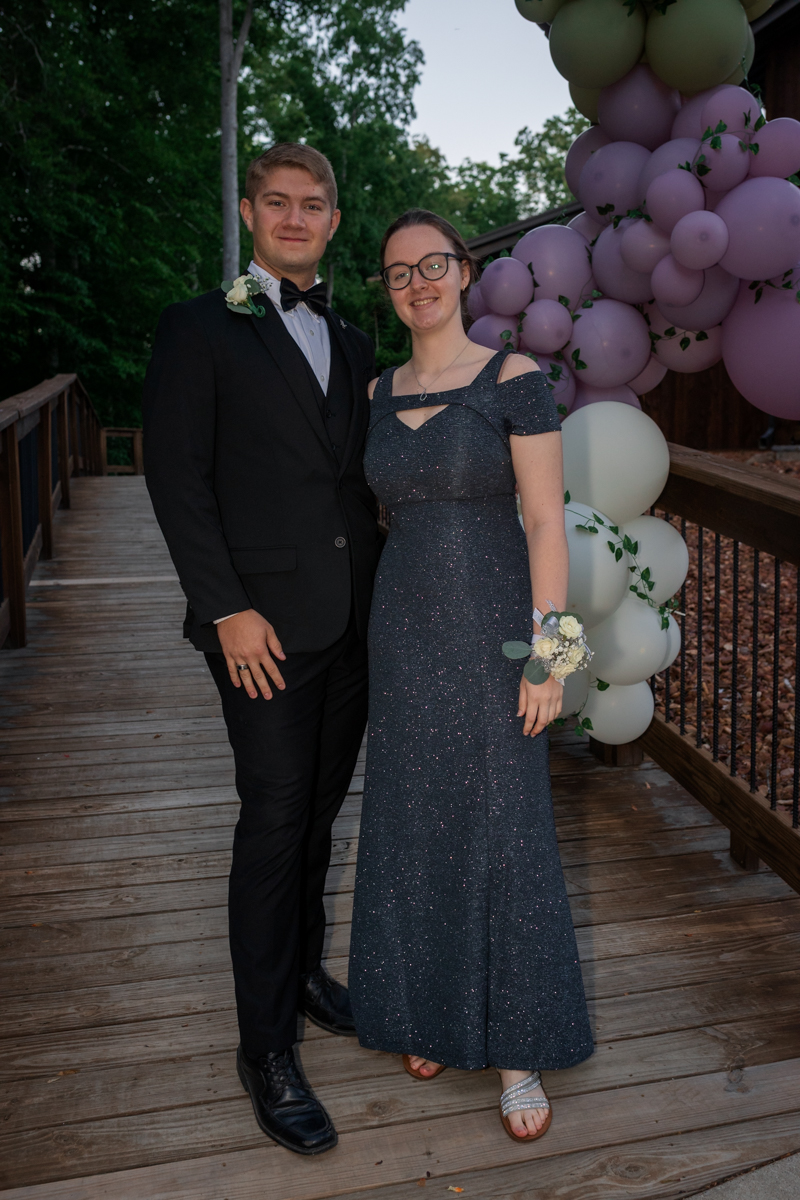 Young couple in formal attire posing on a wooden deck with a lilac and white balloon arch in the background.