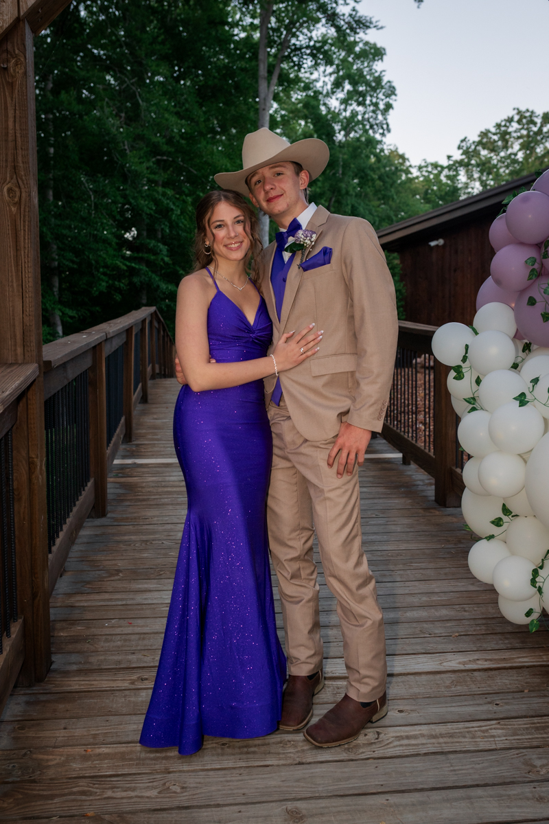 Couple posing on a wooden bridge at a formal event: woman in a purple glitter gown and man in a beige suit with a cowboy hat, purple accents, balloons nearby.